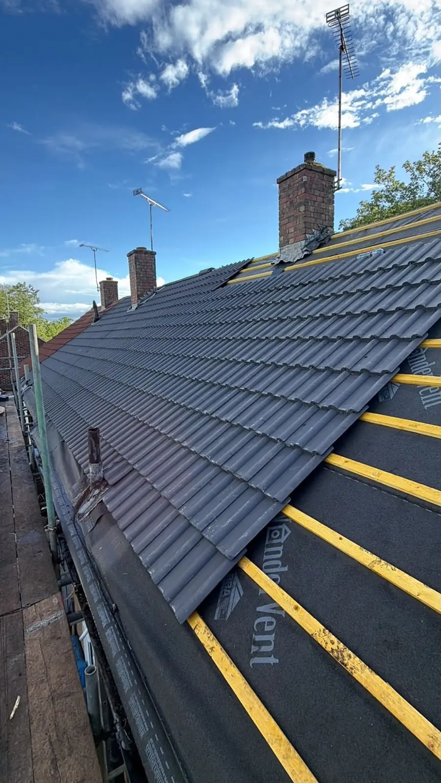 Dark tiles going on over yellow battens and membrane, two chimneys ahead, scaffold below