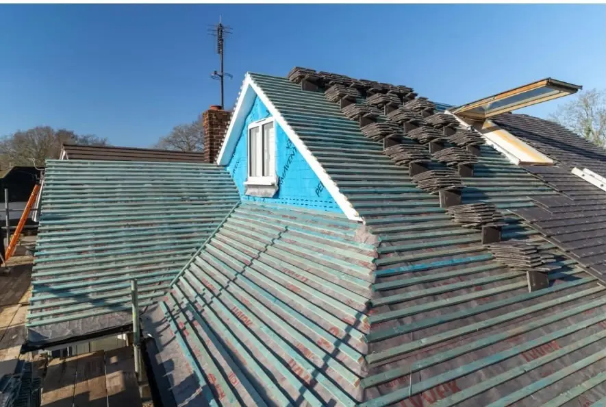 Roof being re-tiled over green membrane and battens beside a blue dormer, old tiles stacked ready to go