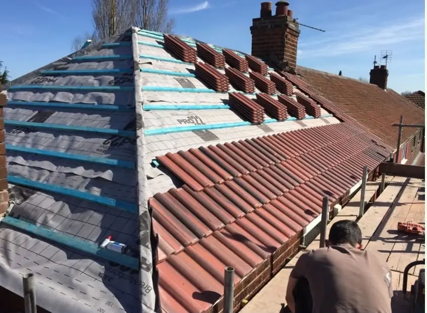 Red tiles being laid over teal battens and membrane, one side done, brick chimney behind
